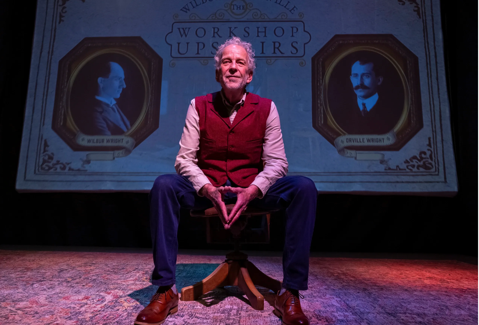Robert Leddy seated onstage between projected portraits of Wilbur and Orville Wright
