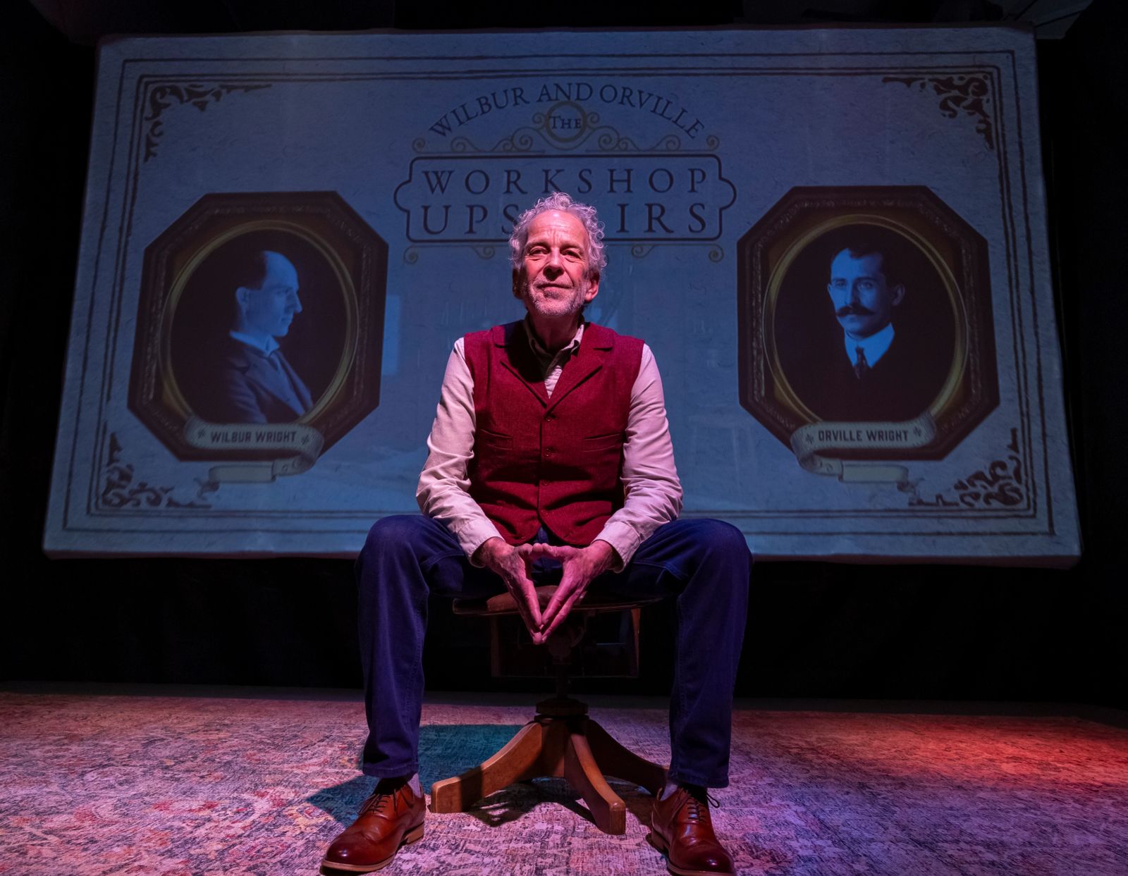 Robert Leddy seated onstage between portraits of Wilbur and Orville Wright